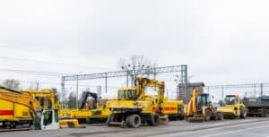 Construction vehicles, such as excavators and bulldozers, are parked along a road near railway tracks. The sky is overcast, and power lines run in the background.