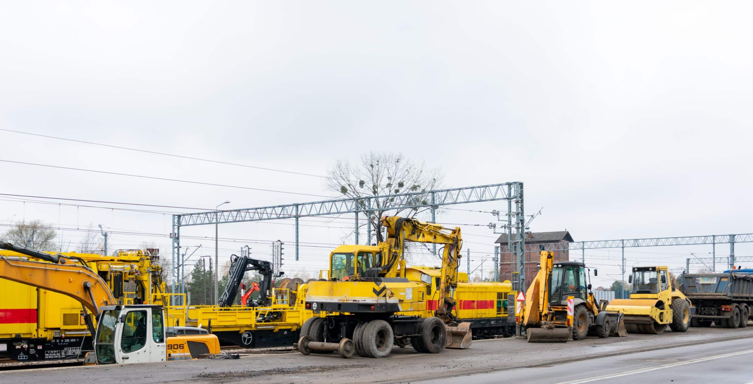 Construction vehicles, such as excavators and bulldozers, are parked along a road near railway tracks. The sky is overcast, and power lines run in the background.