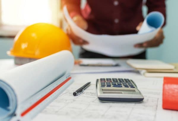 A person examines rolled blueprints on a desk with a calculator, pen, and orange hard hat nearby, suggesting an office or construction planning environment.