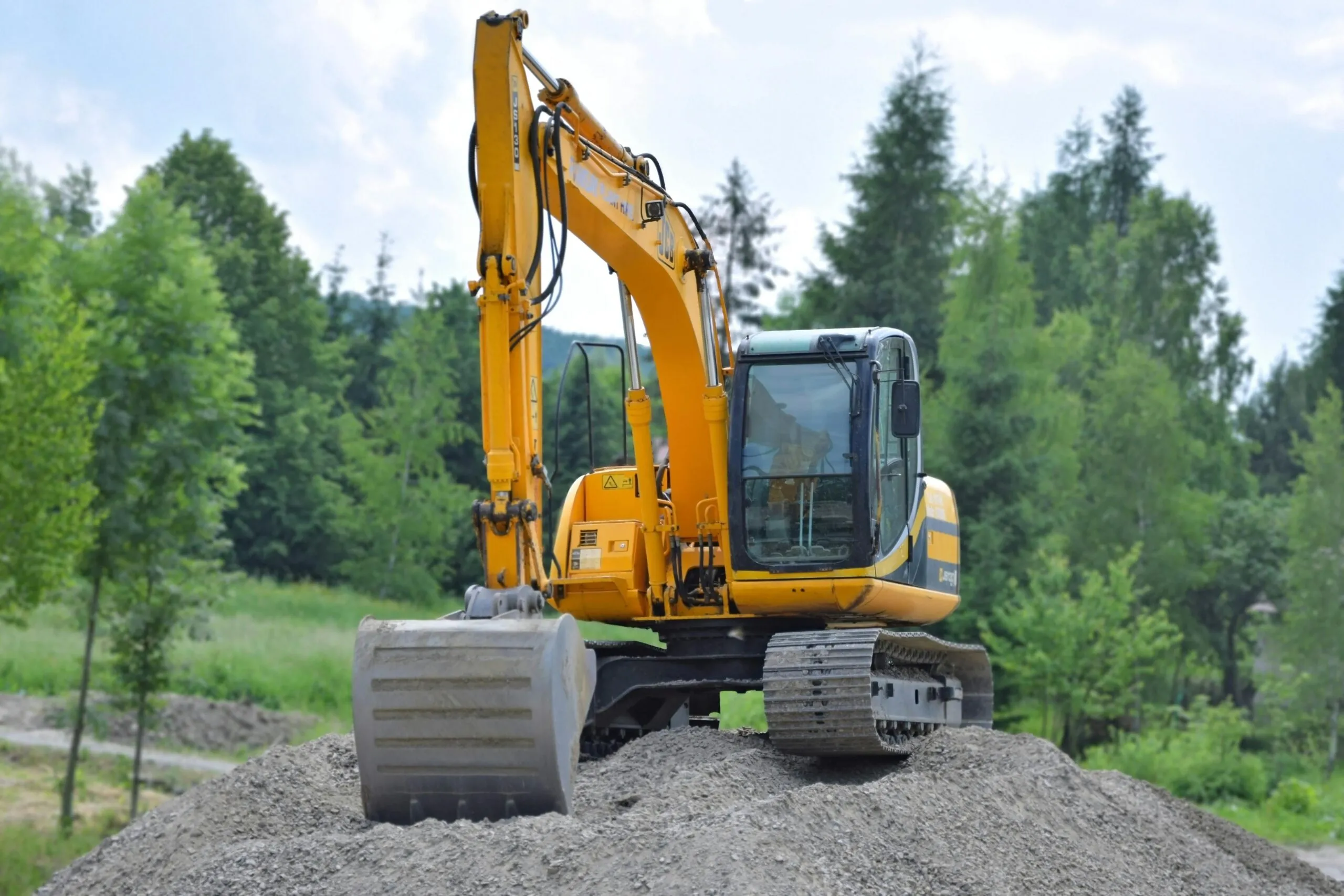 A yellow excavator sits on a gravel mound within a lush, forested area, surrounded by trees and partly cloudy skies.