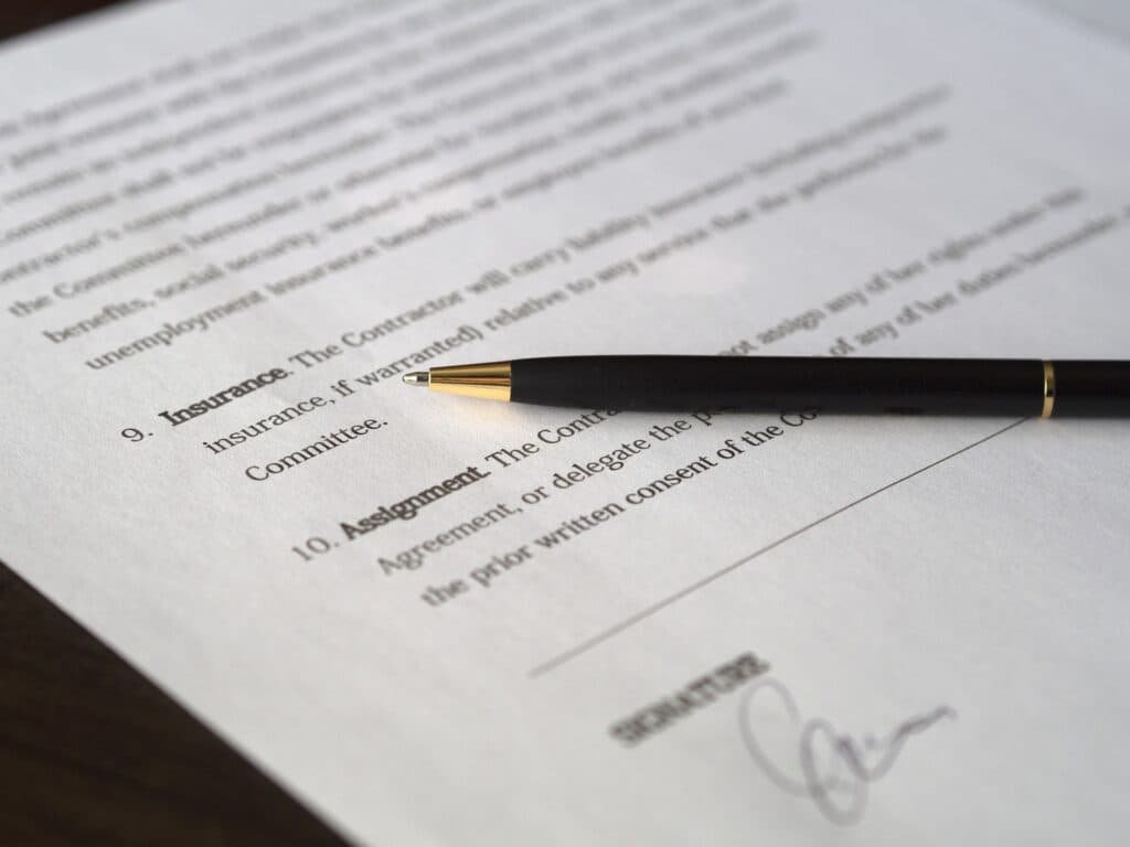A black pen rests on a document with numbered sections titled "Insurance" and "Assignment." There's a signature near the bottom, indicating the document's potential legal or formal nature.
