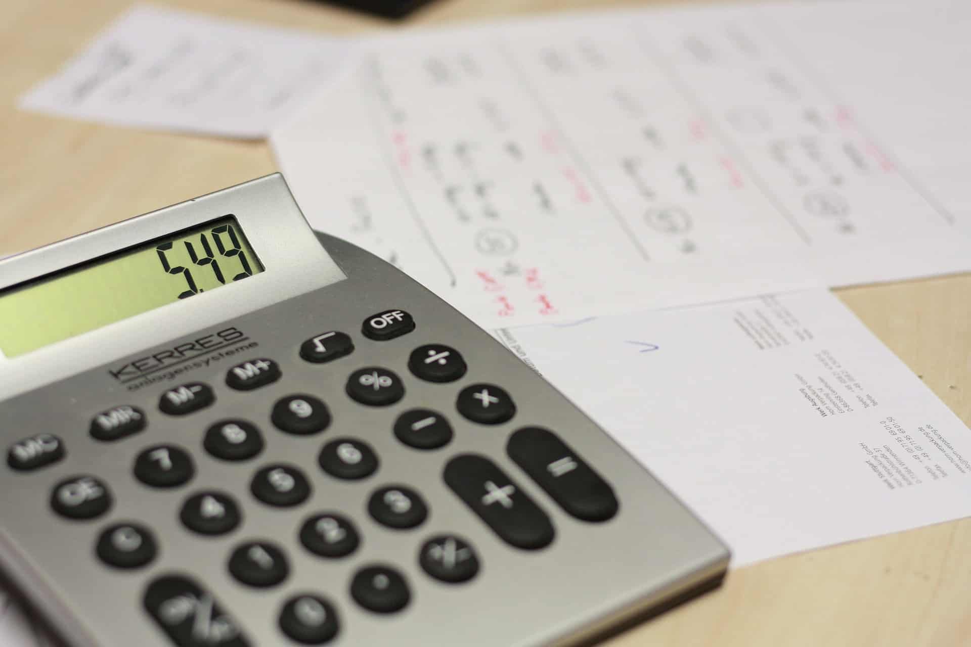 A calculator displaying "5.49" rests on a wooden surface with a few sheets of paper scattered around, suggesting a work or study environment.