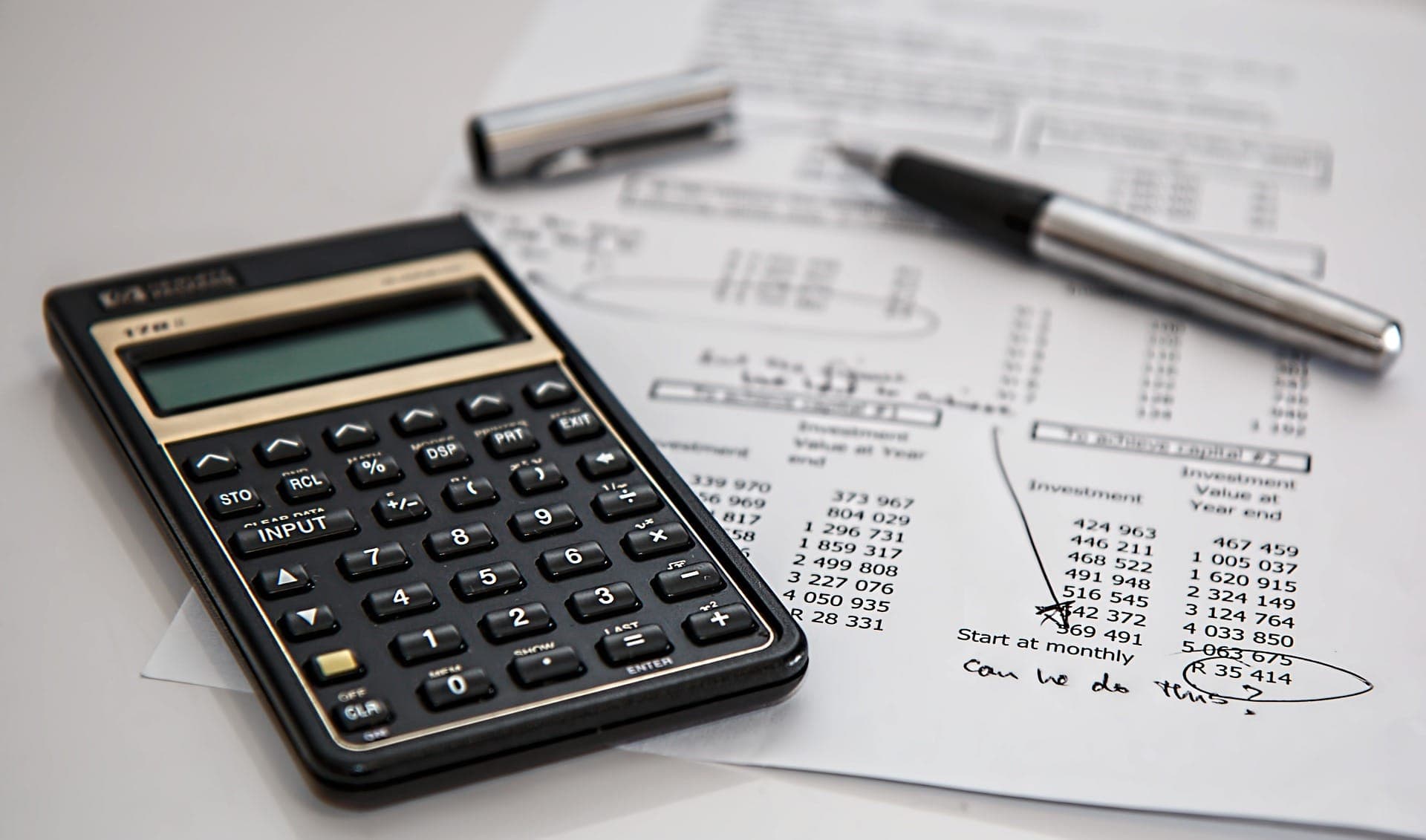 A calculator sits on documents with numerical data and calculations, accompanied by an uncapped pen. The context suggests a financial or mathematical setting, focused on analysis or budgeting tasks.