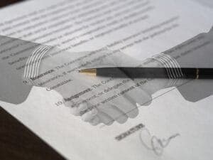 A pen overlays a handshake on a blurred contract. The document includes text about "The Ownership" and "Assignment," with a signature at the bottom, implying a finalized agreement.