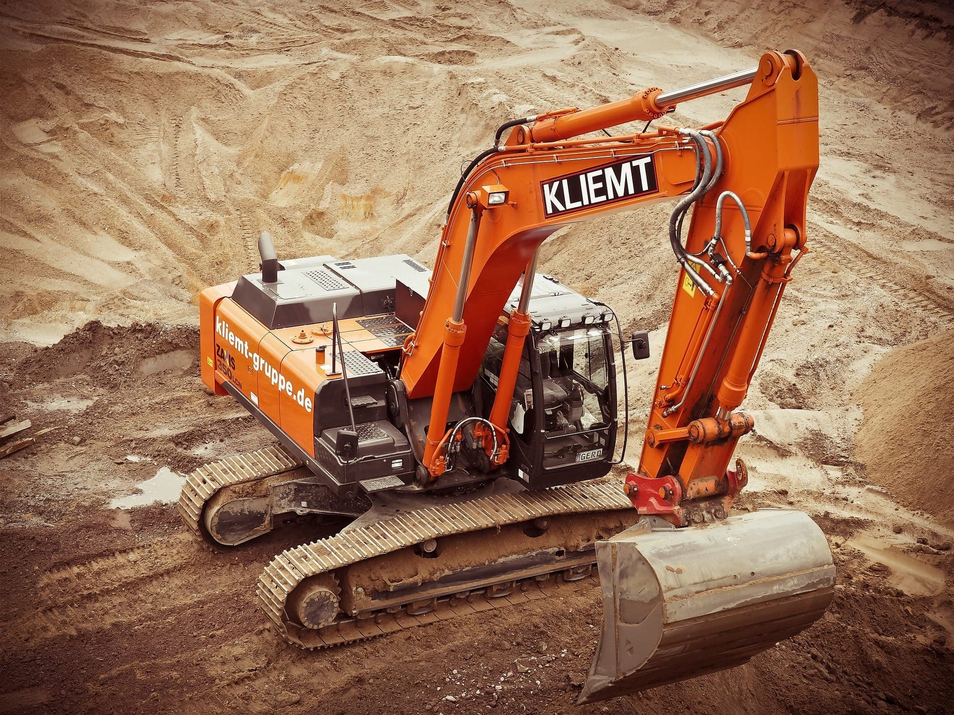 An orange excavator labeled "KLIEMT" with a bucket is digging at a construction site, surrounded by dirt and earthmoving activity.
