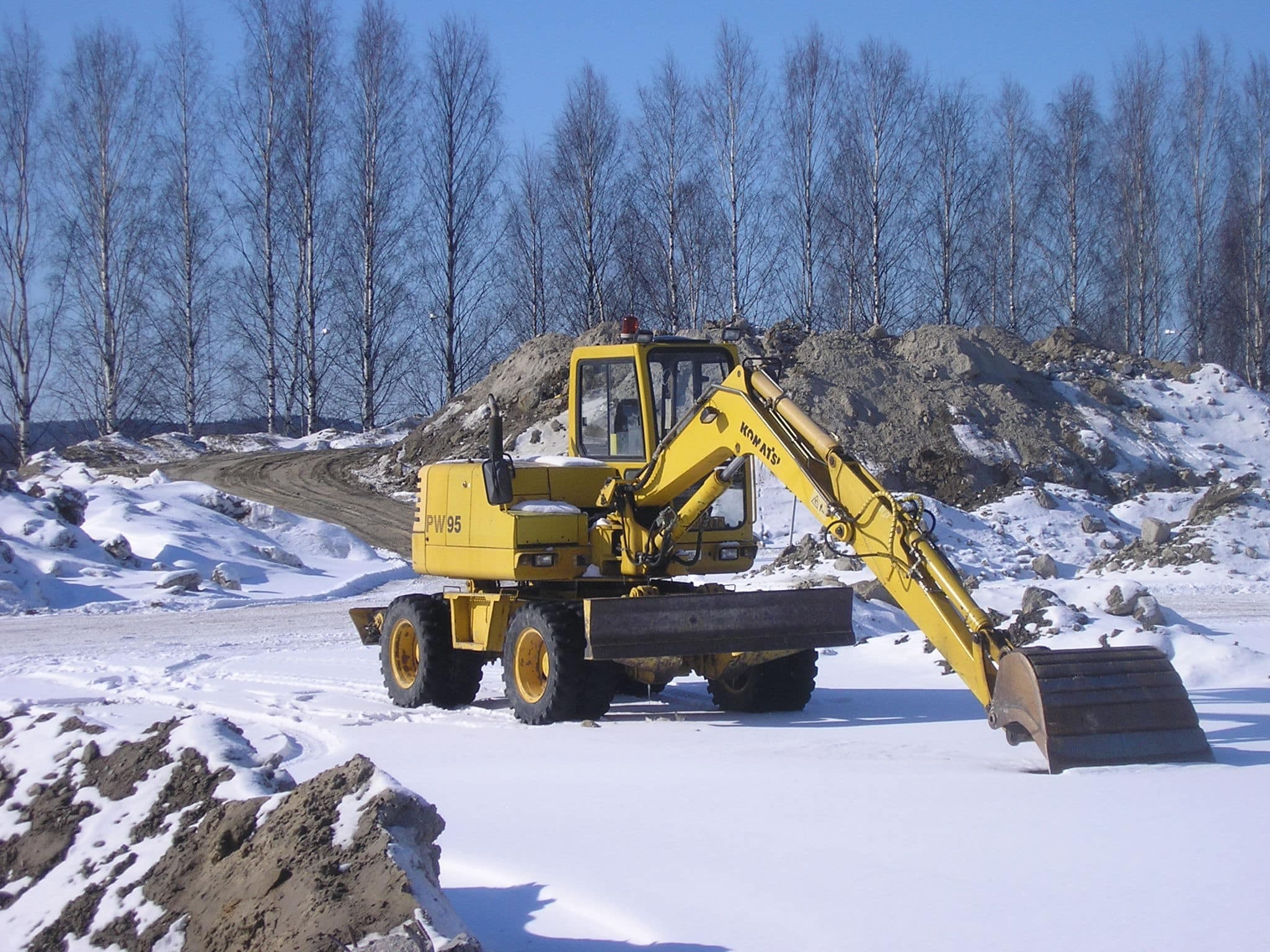 A yellow excavator rests on snow-covered ground, surrounded by mounds of snow and dirt, with leafless trees in the background under a clear blue sky.