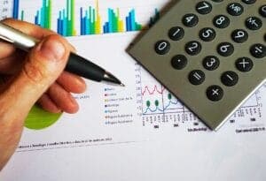 A hand holds a pen hovering over financial documents with colorful bar and line graphs, next to a calculator. The context includes data analysis or financial review.
