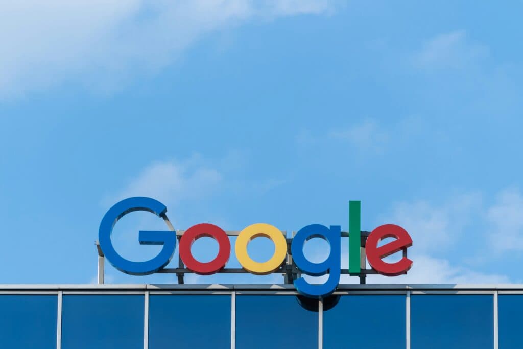 The Google logo appears on a glass building facade under a clear blue sky, featuring large, colorful letters in green, red, yellow, and blue.