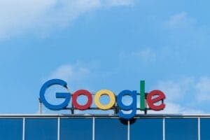 The Google logo appears on a glass building facade under a clear blue sky, featuring large, colorful letters in green, red, yellow, and blue.
