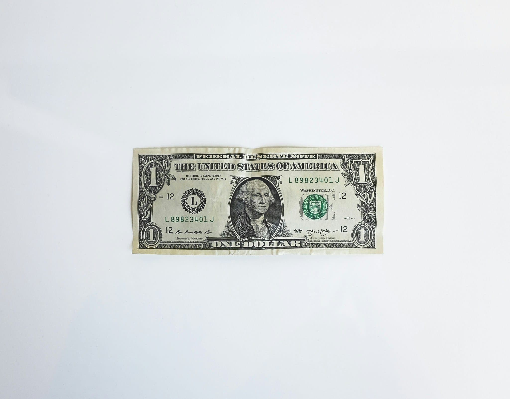 A U.S. one-dollar bill lies flat on a plain white surface. The bill prominently displays George Washington's portrait, with text reading "THE UNITED STATES OF AMERICA" and "ONE DOLLAR."
