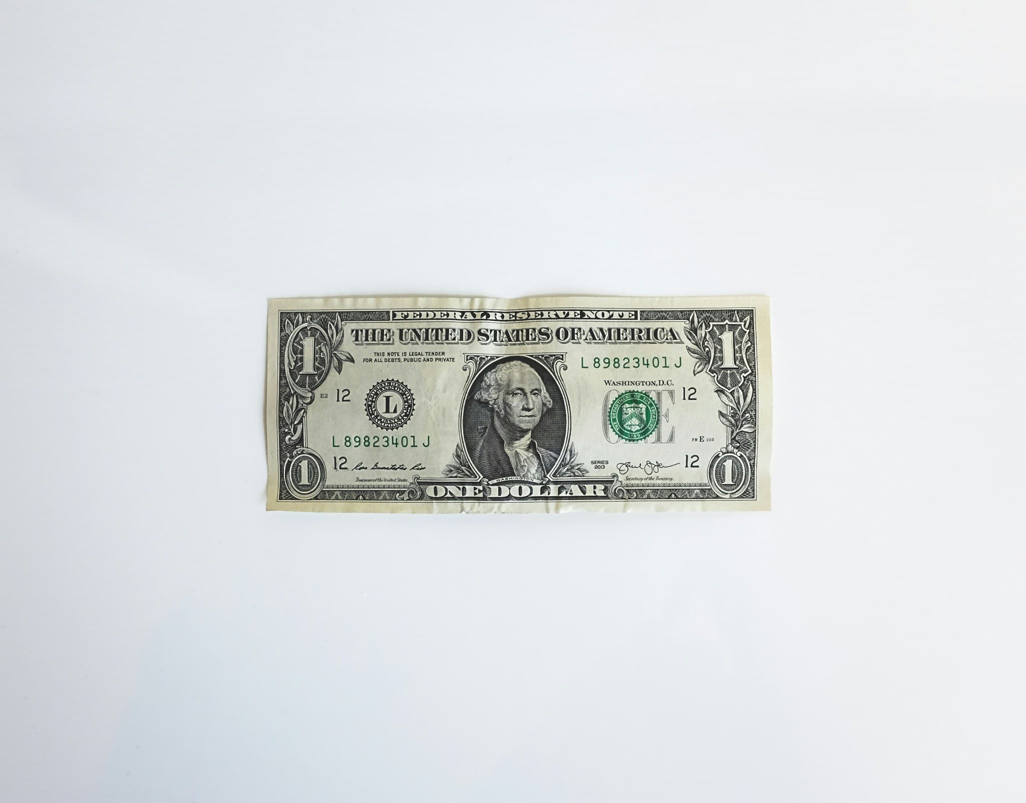 A U.S. one-dollar bill lies flat on a plain white surface. The bill prominently displays George Washington's portrait, with text reading "THE UNITED STATES OF AMERICA" and "ONE DOLLAR."
