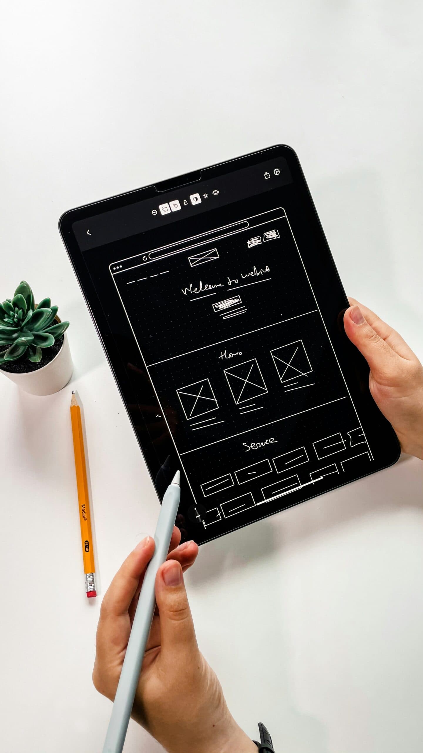A person uses a stylus on a tablet displaying a wireframe design titled "Website Architect." A pencil and a small plant sit on the white surface beside them.