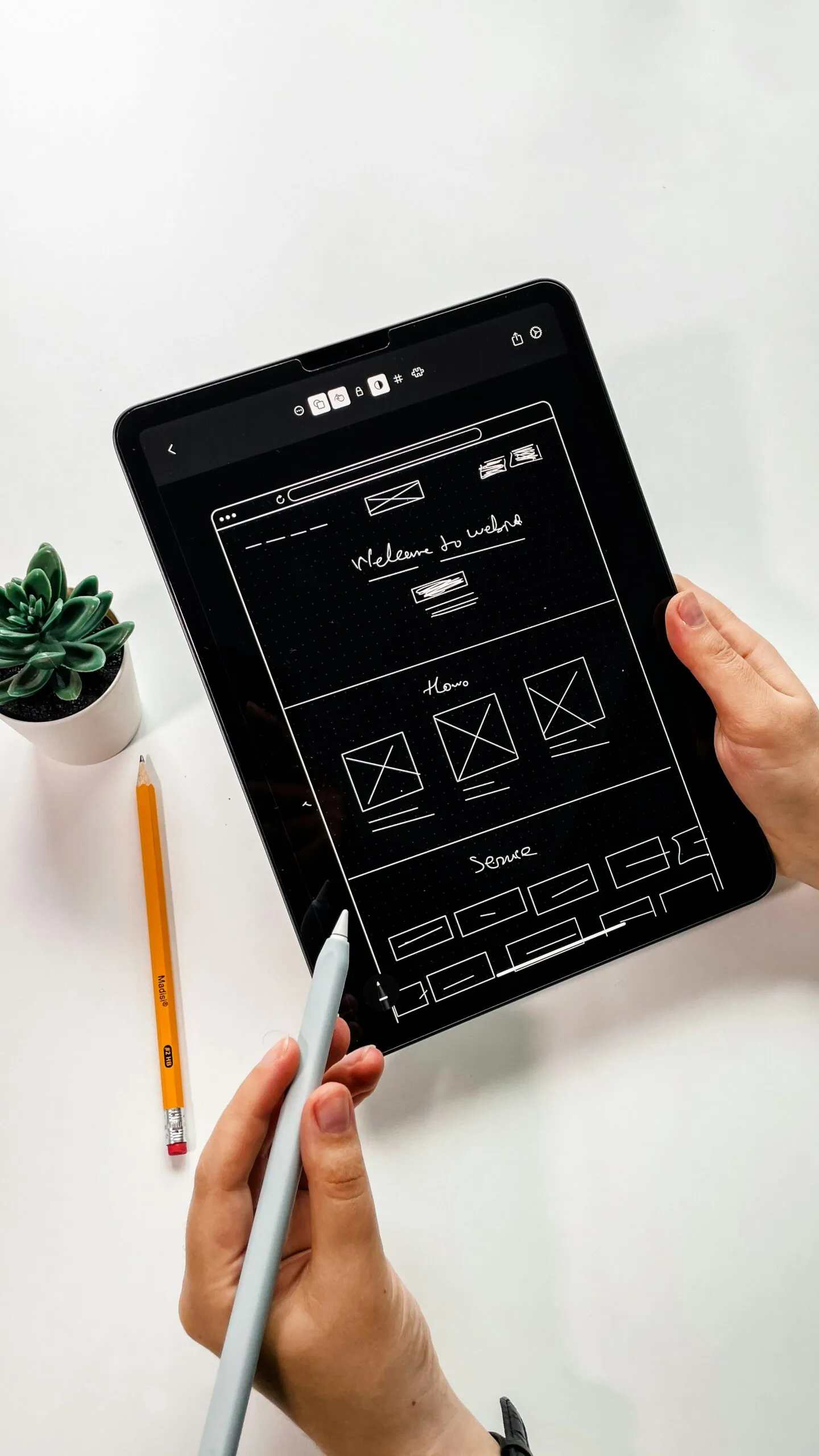A person uses a stylus on a tablet displaying a wireframe design titled "Website Architect." A pencil and a small plant sit on the white surface beside them.