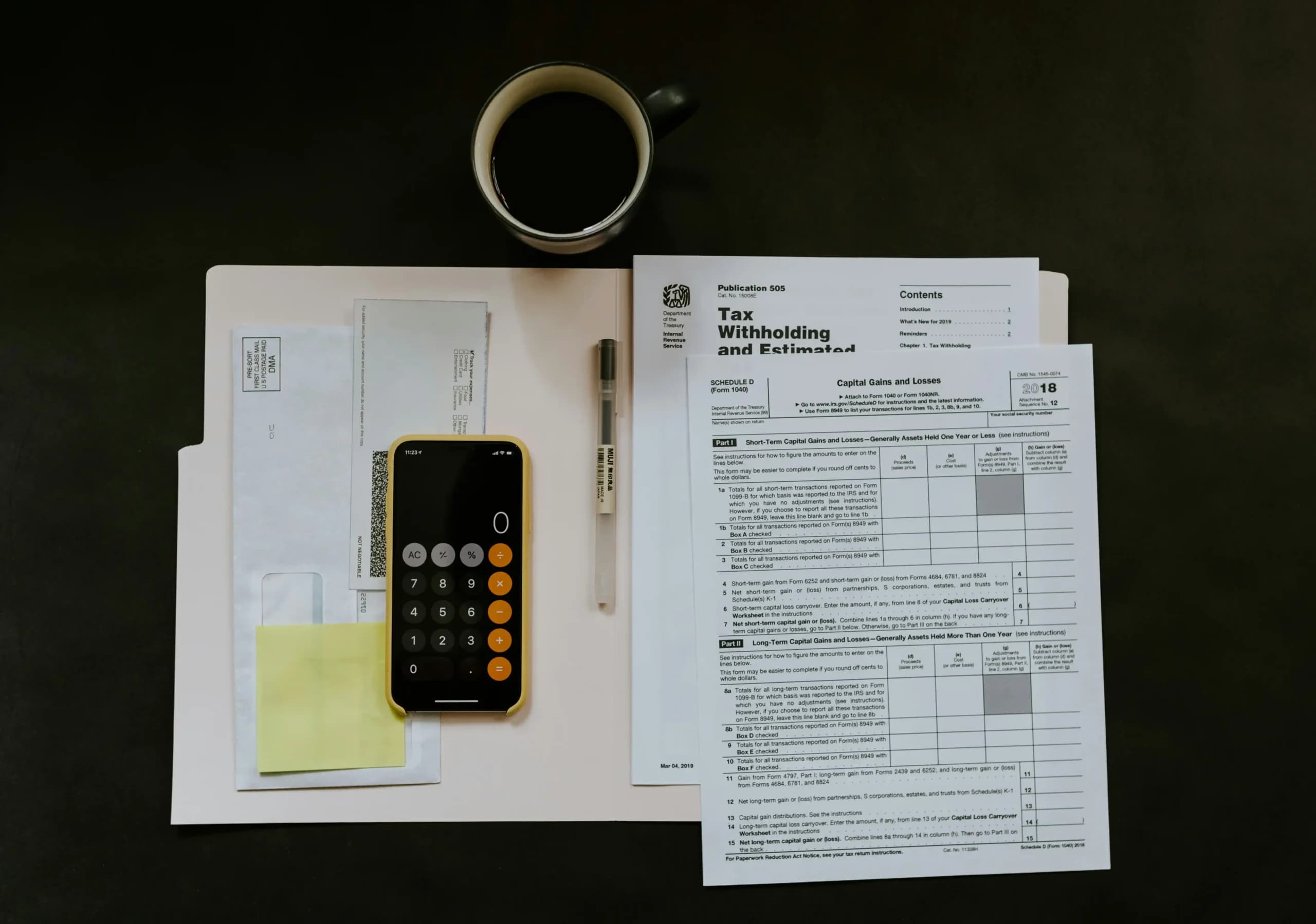 A smartphone with a calculator app lays on letters and sticky notes; beside it are IRS tax documents with a pen, and a mug of black coffee on a dark desk.
