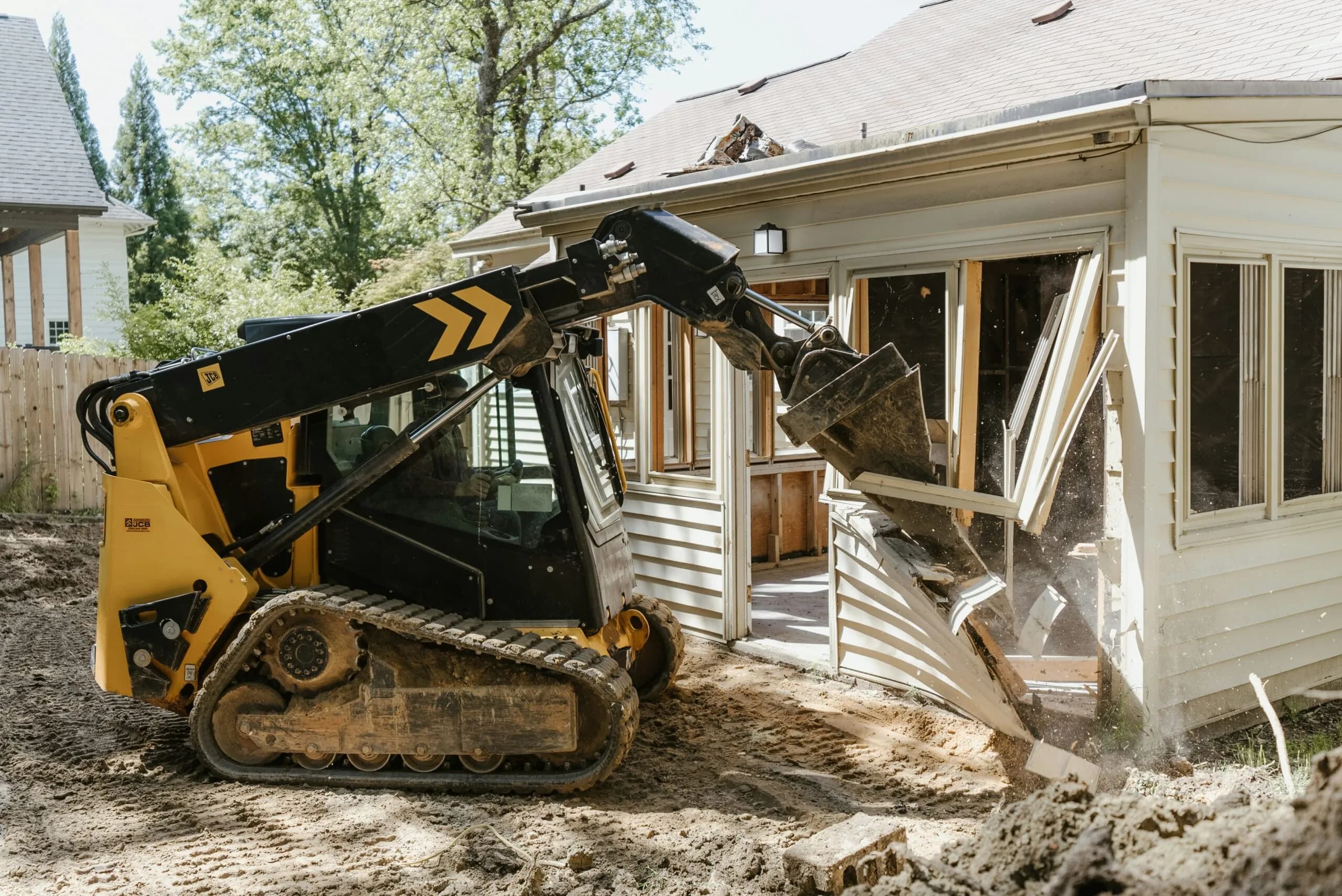 A small excavator demolishes a section of a house wall using its bucket in a residential area, surrounded by trees and clear skies.