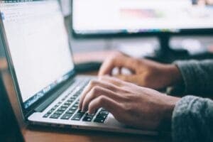 Hands typing on a laptop keyboard, open to a text document. The background shows blurred computer screens, suggesting a work or study environment.