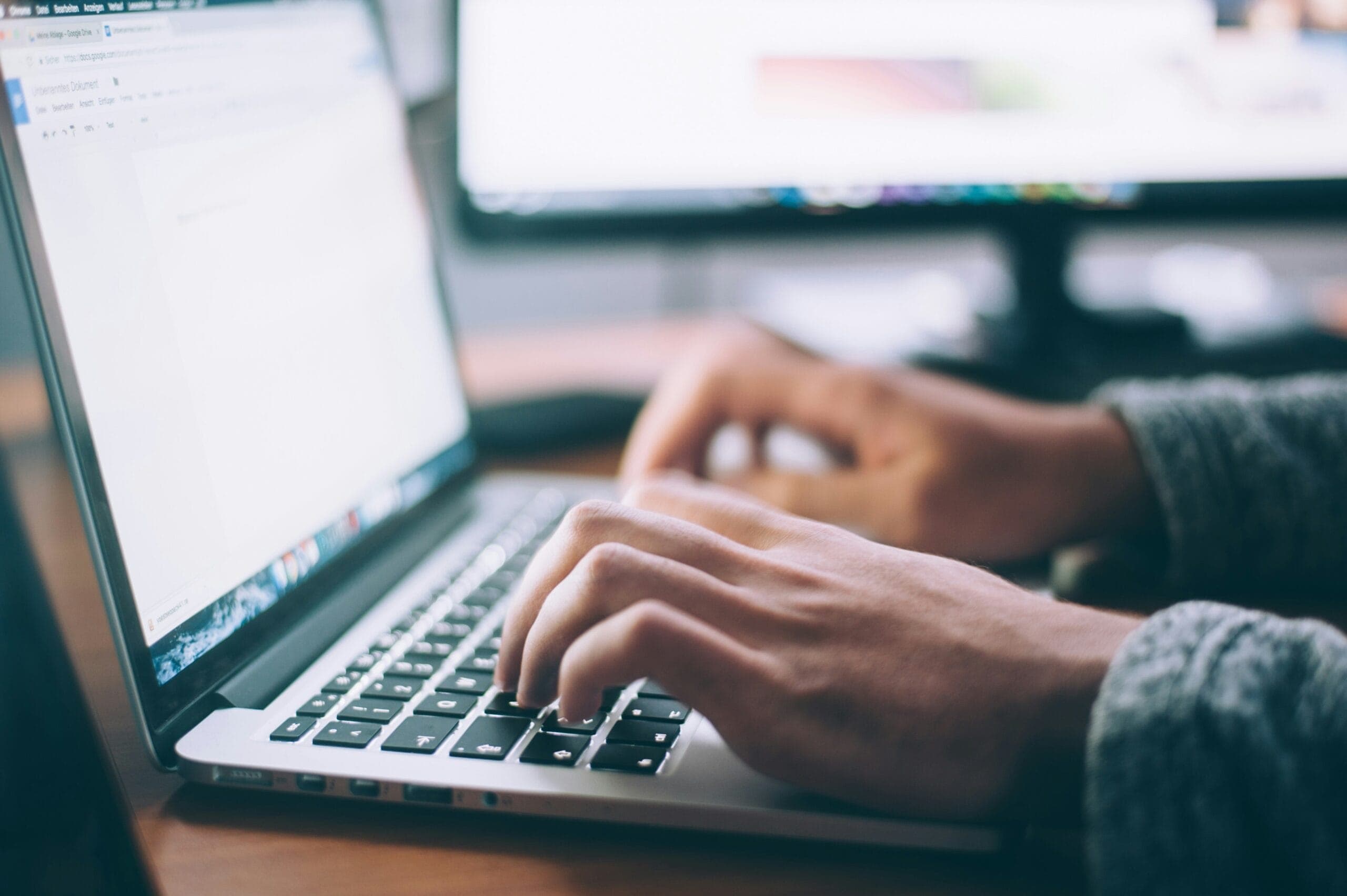 Hands typing on a laptop keyboard, open to a text document. The background shows blurred computer screens, suggesting a work or study environment.