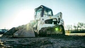 A skid steer loader rests on a construction site, bathed in sunlight, surrounded by dirt and a sparse line of trees in the background.