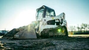 A skid steer loader rests on a construction site, bathed in sunlight, surrounded by dirt and a sparse line of trees in the background.