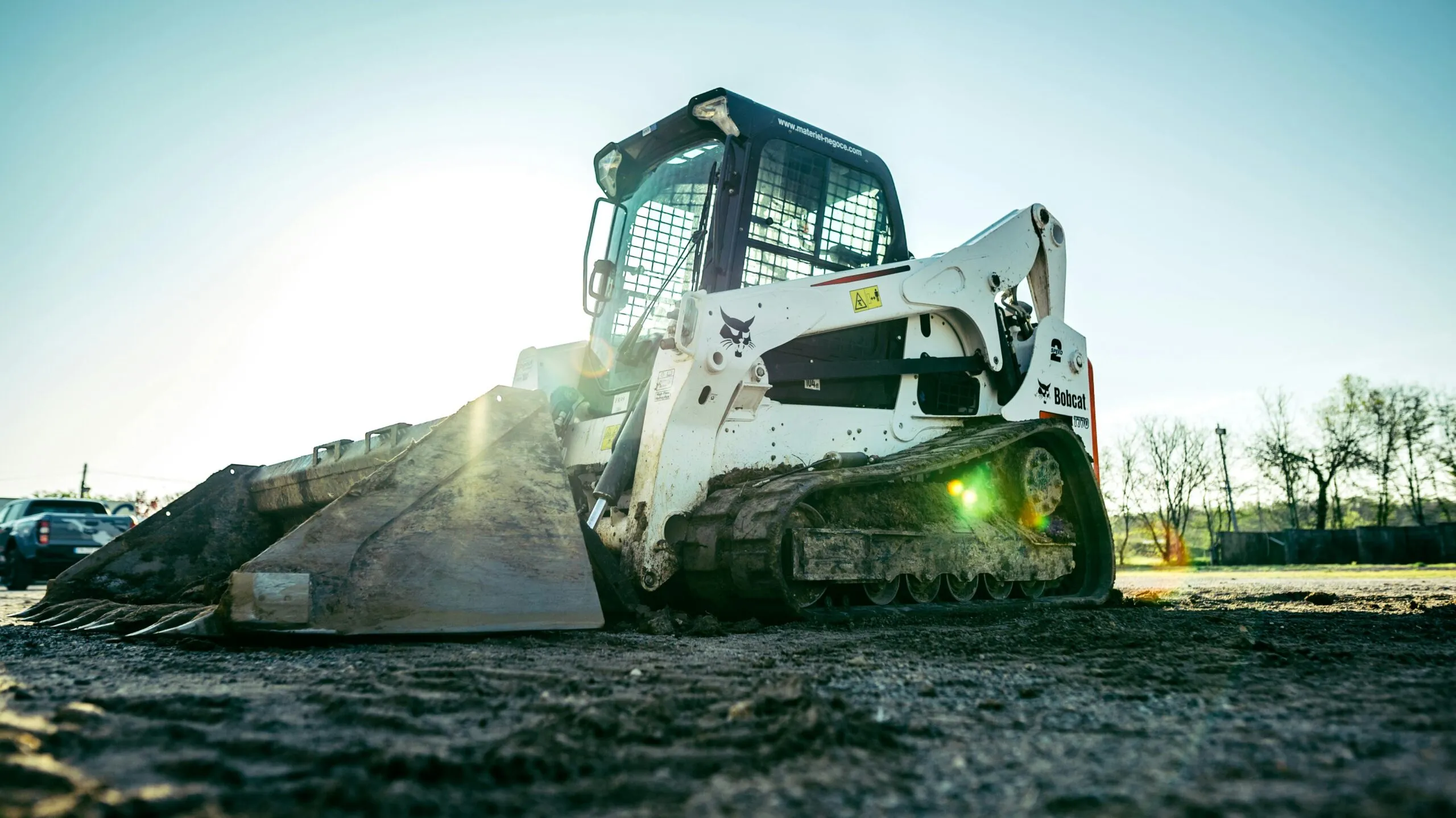 A skid steer loader on a construction site, surrounded by dirt with a sparse line of trees behind.